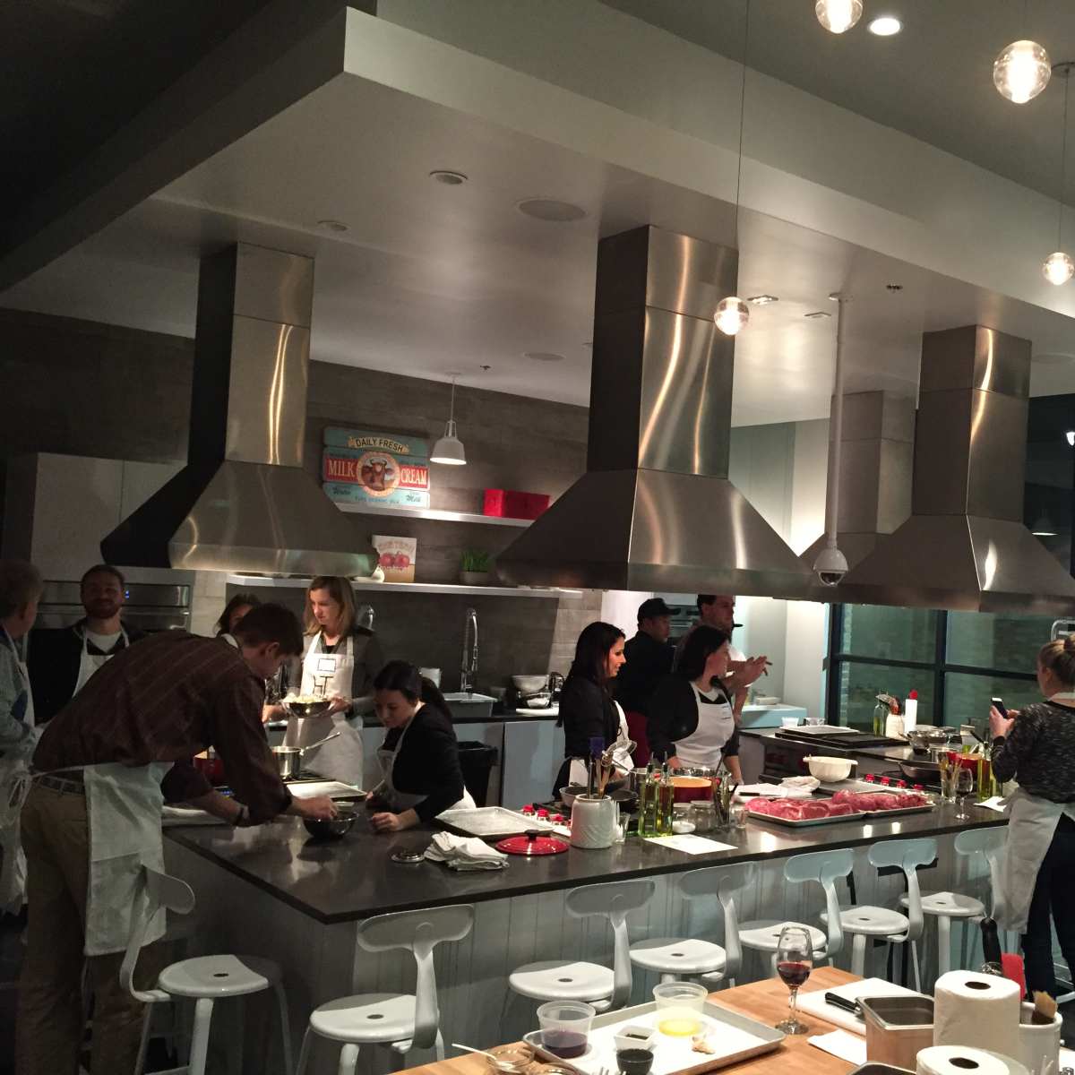 A group of people are cooking together in a modern kitchen, equipped with stainless steel appliances and large cooking surfaces.