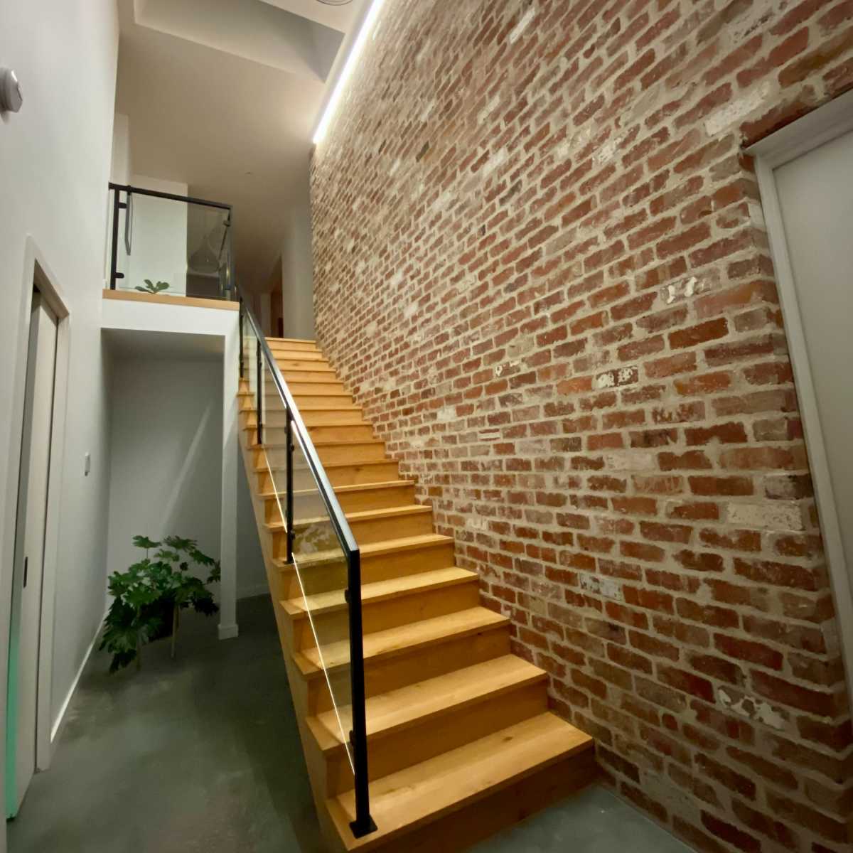 A wooden stairway leading to an upper level, flanked by a brick wall and illuminated by pendant lights and recessed lighting.