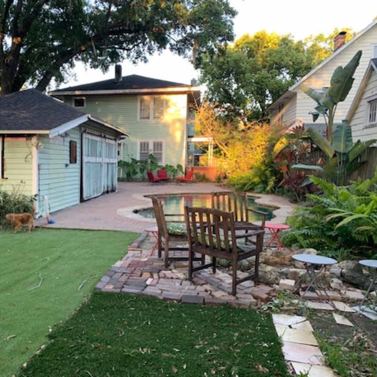 A backyard with a small pool, a patio area with chairs, a green lawn, and two houses in the background.