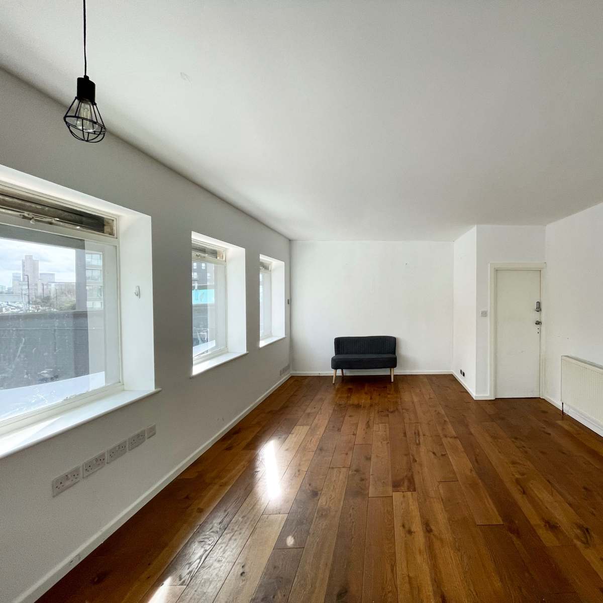 A spacious, empty room with wooden flooring, a single gray sofa, large windows, and a door leading to another area.