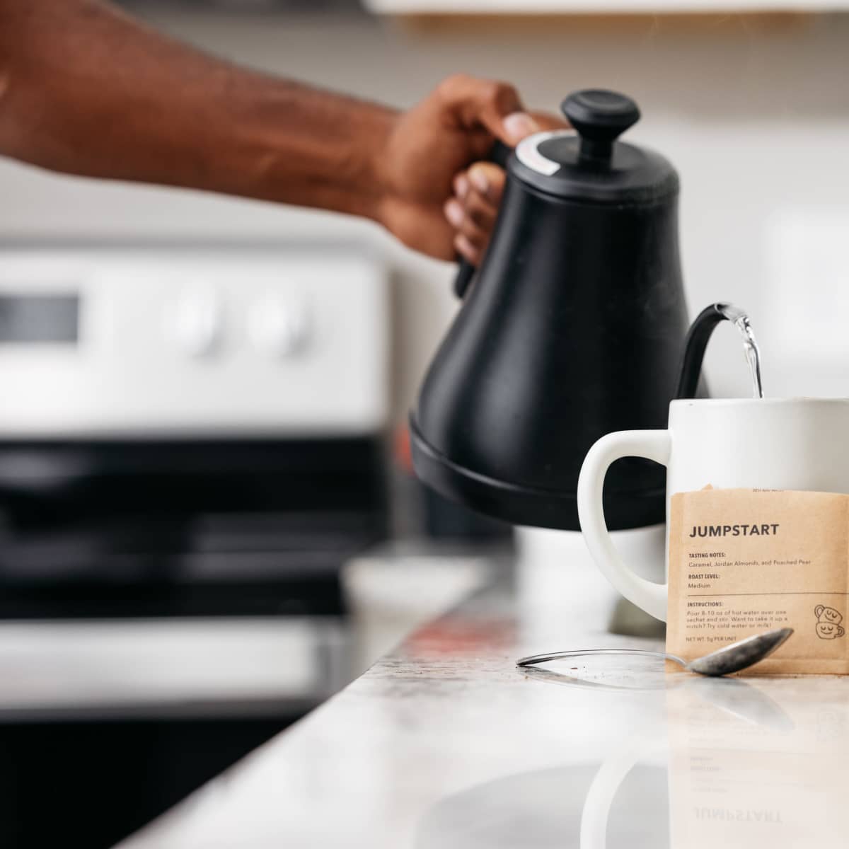 A person pours hot water from a kettle into a mug with a tea bag labeled "JUMPSTART" on a kitchen countertop.