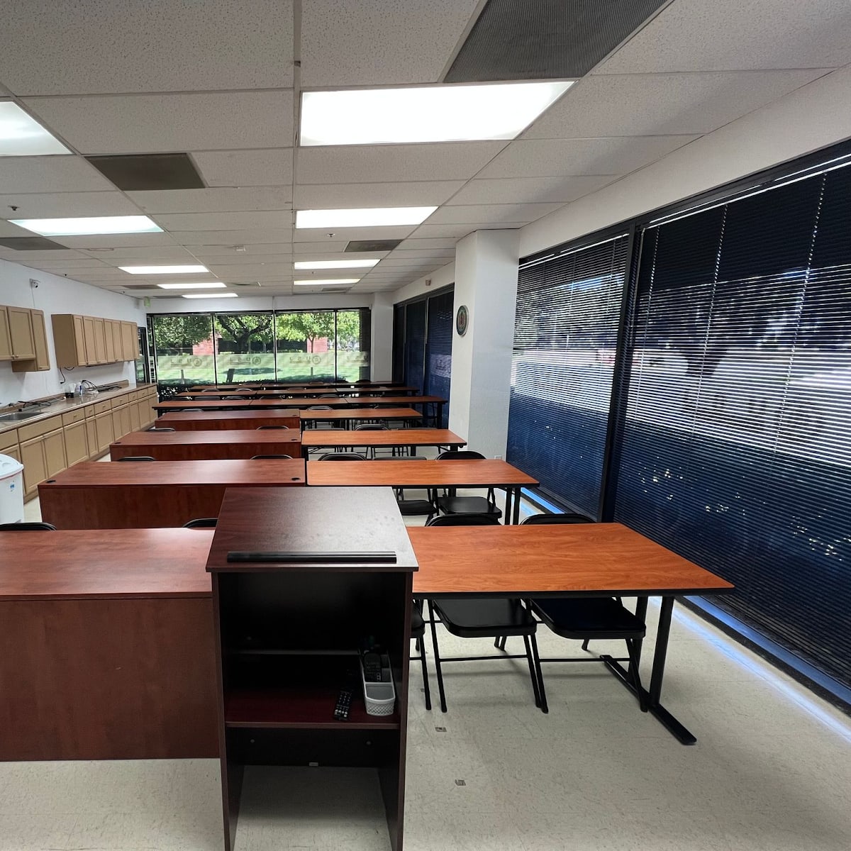 A classroom setup with rows of wooden desks and a kitchen area in the background, illuminated by natural light from large windows.
