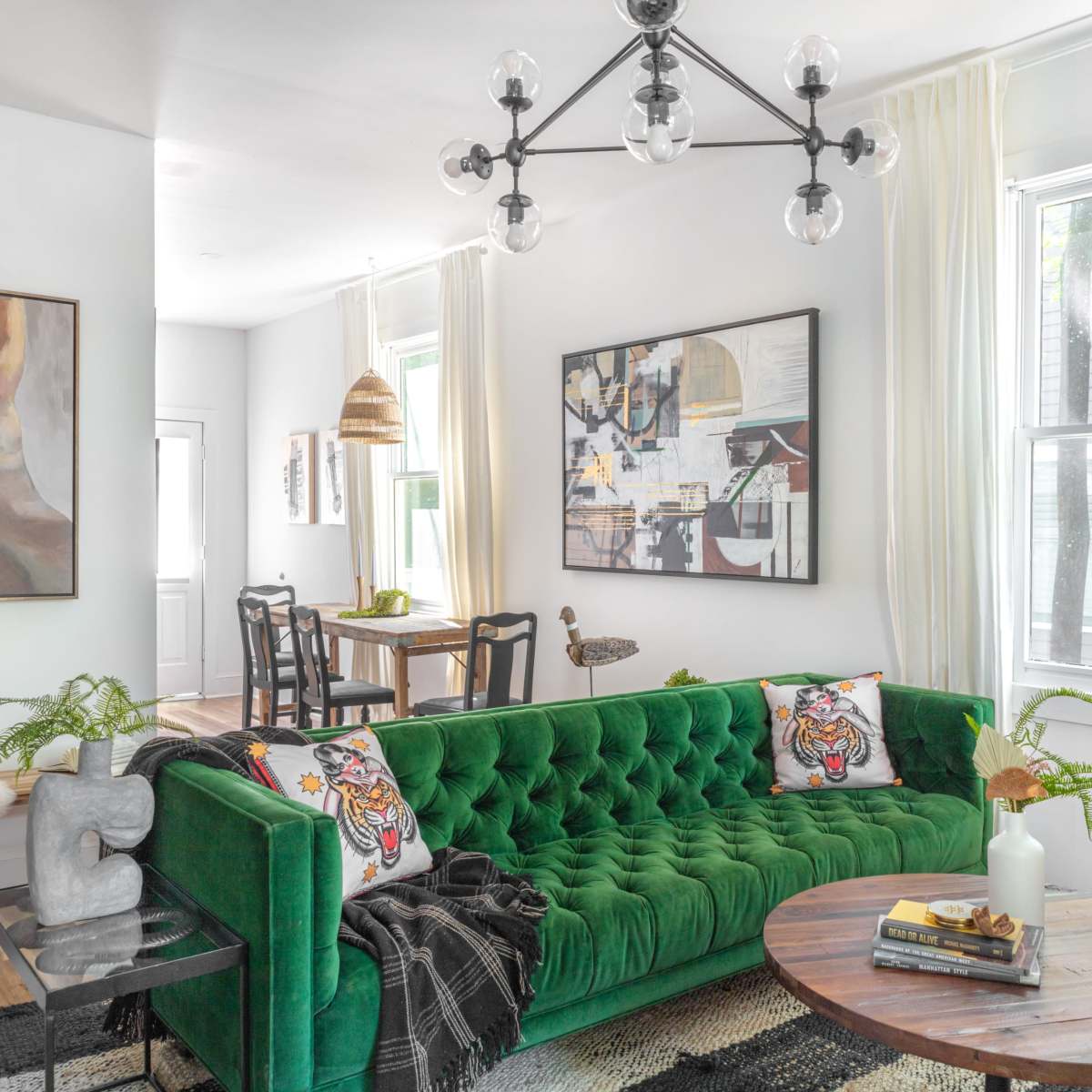 A modern living area featuring a green velvet sofa with decorative pillows, a round wooden coffee table, and large windows allowing natural light to fill the space.