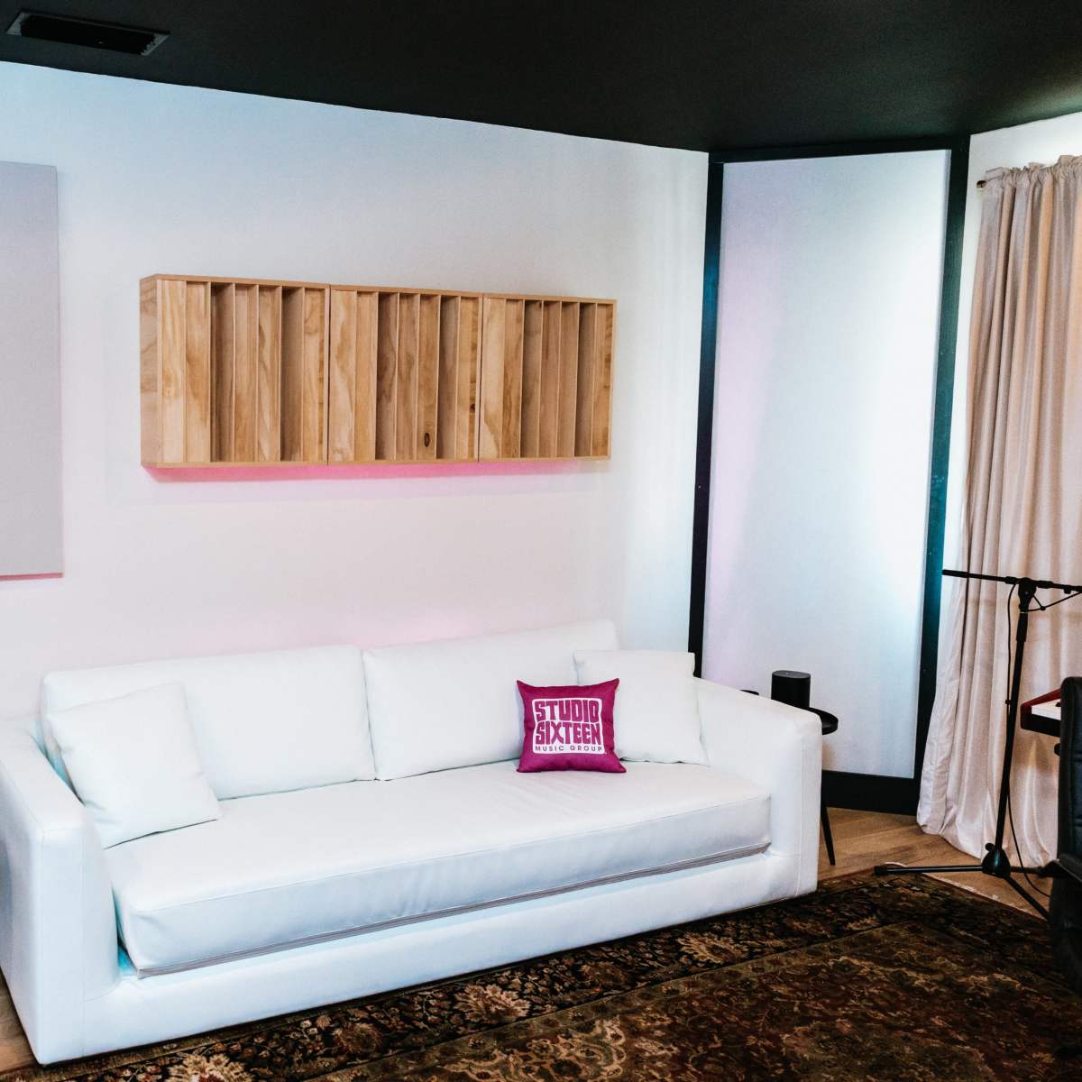 A minimalist room featuring a white couch with a pink decorative pillow, a black chair, a small round table, wooden shelving, and a large soundproof panel on the wall.