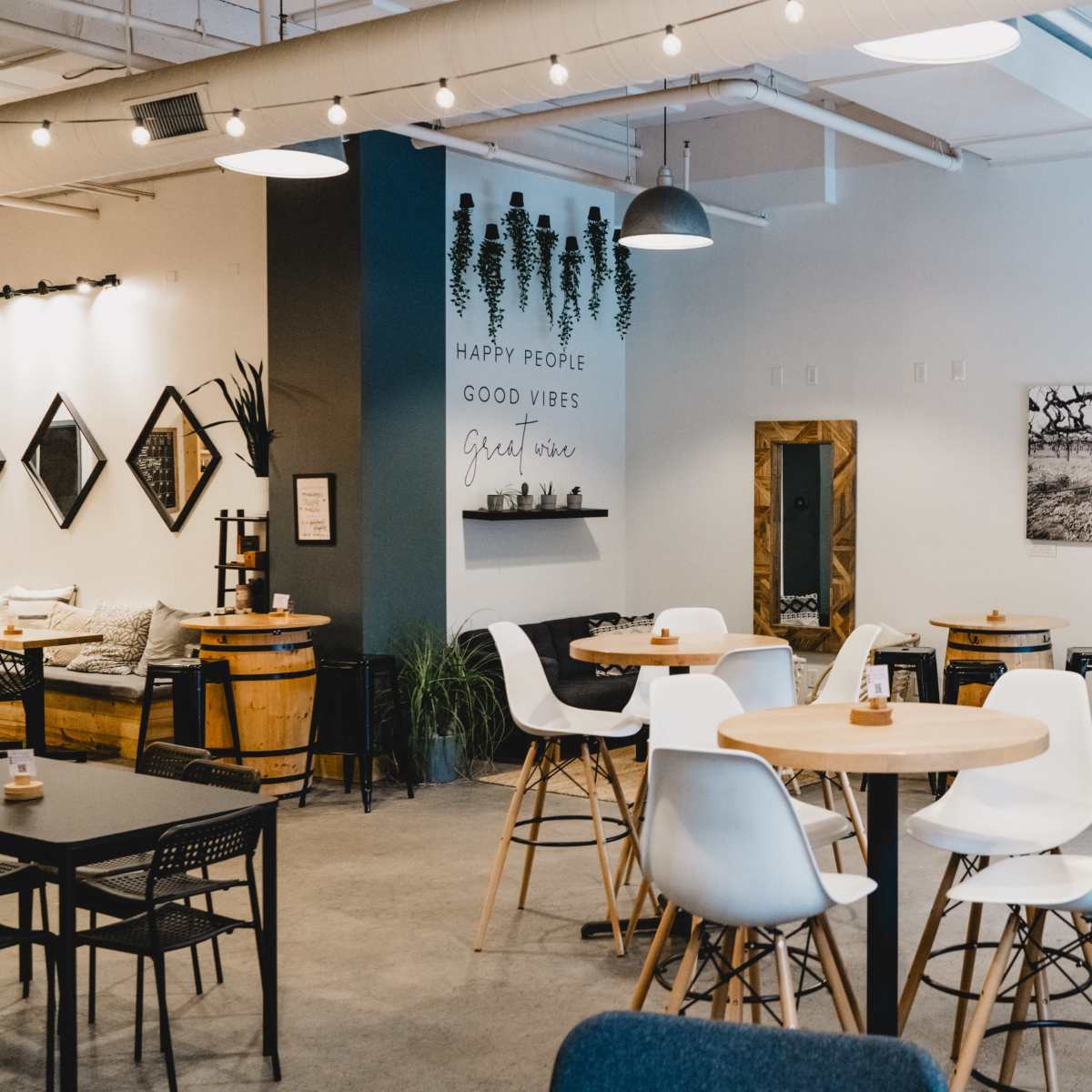 A modern café interior with round wooden tables, white chairs, and decorative plants, featuring a wall with inspirational phrases and black-and-white photographs.