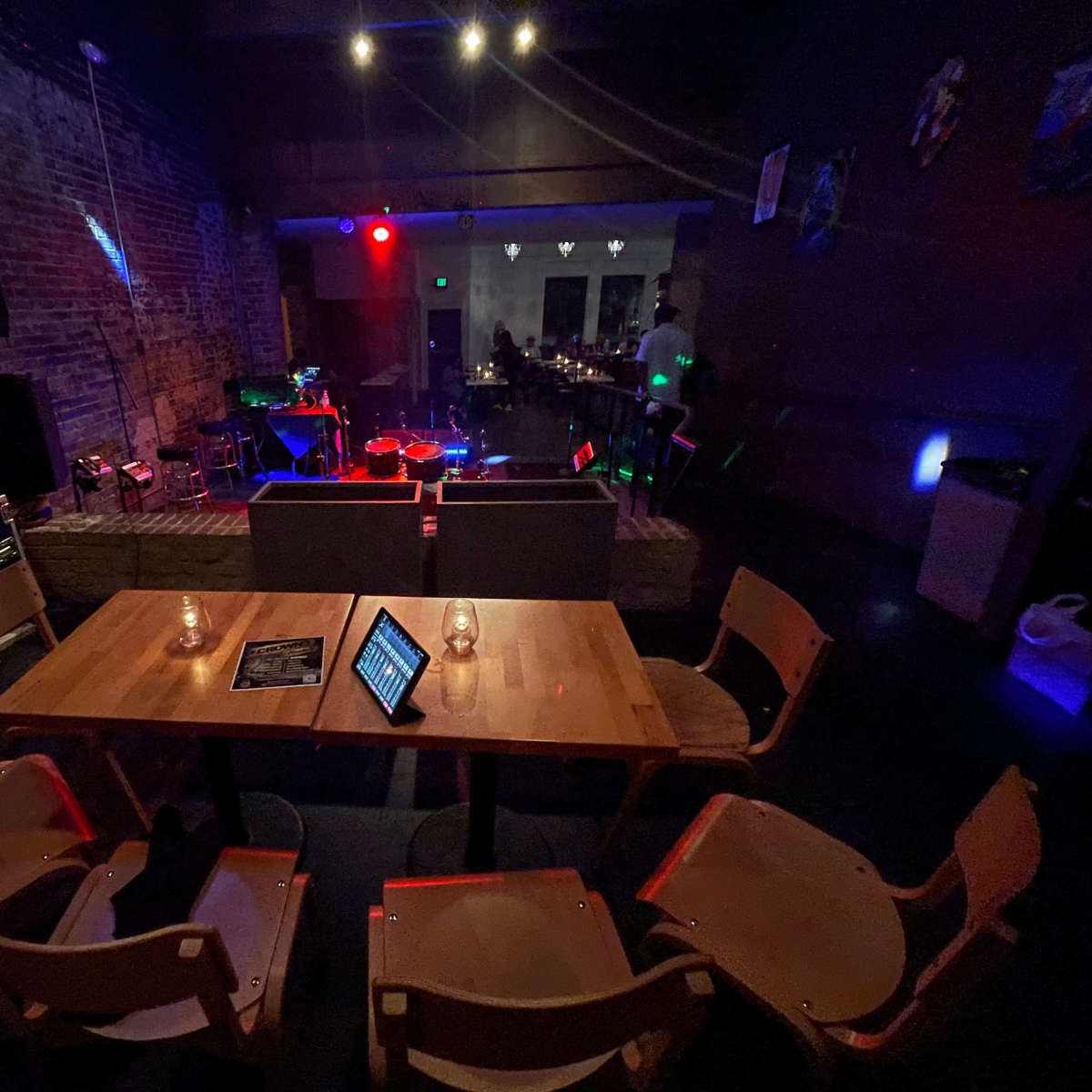 An empty restaurant or bar setting with wooden tables and chairs, soft lighting, and a stage area in the background.
