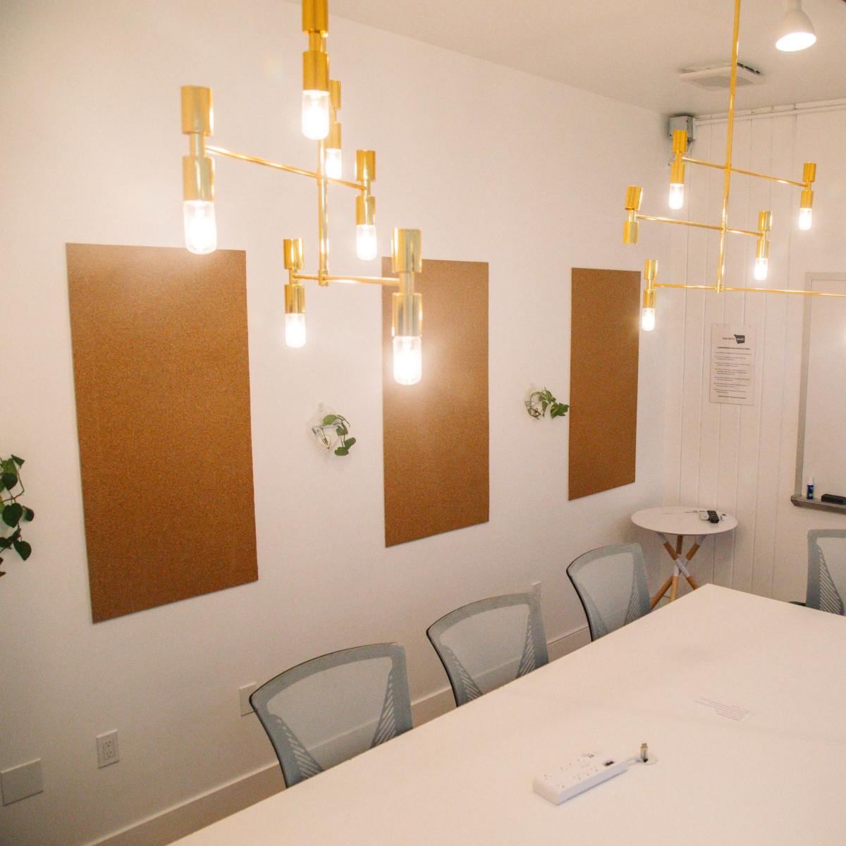 A modern conference room features a long white table, gold light fixtures, corkboard panels on the walls, and a whiteboard at one end.