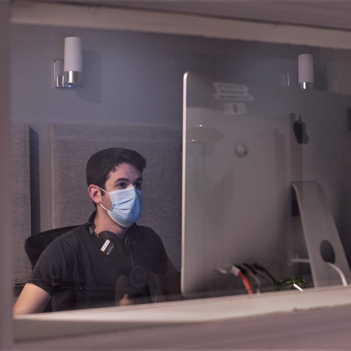 A young man wearing a face mask sits at a desk in front of a computer, with headphones around his neck, in a room visible through a window.