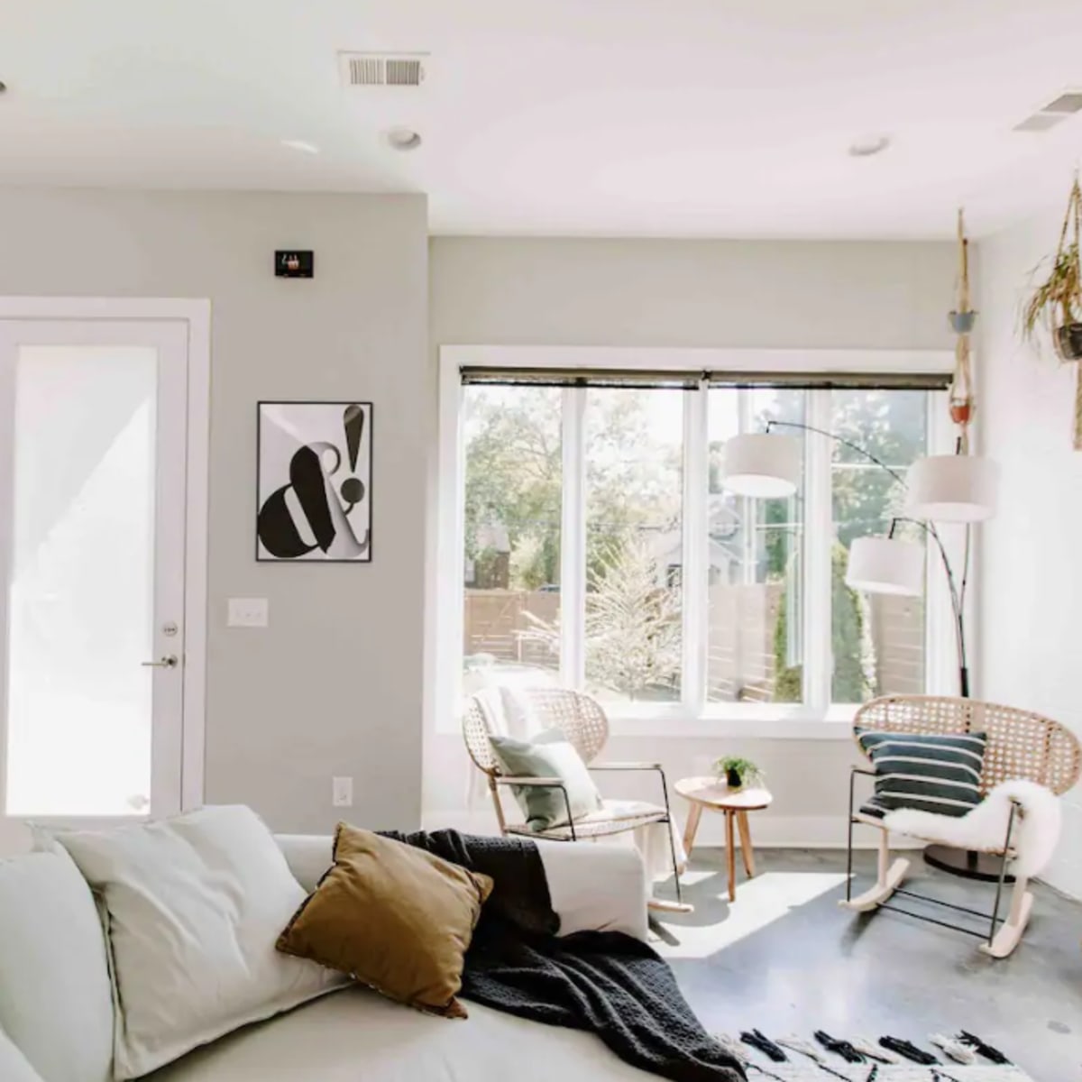 A bright, modern living room with a white sofa, a framed artwork on the wall, and a sunlit corner featuring two chairs and a large window.