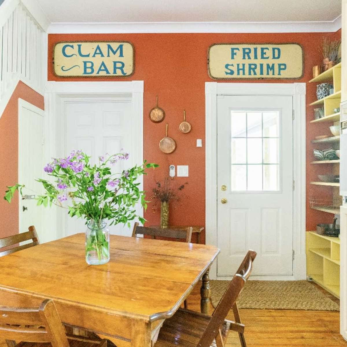 A wooden dining table is surrounded by chairs in a room with orange walls, featuring signs labeled "CLAM BAR" and "FRIED SHRIMP" above the doorway.