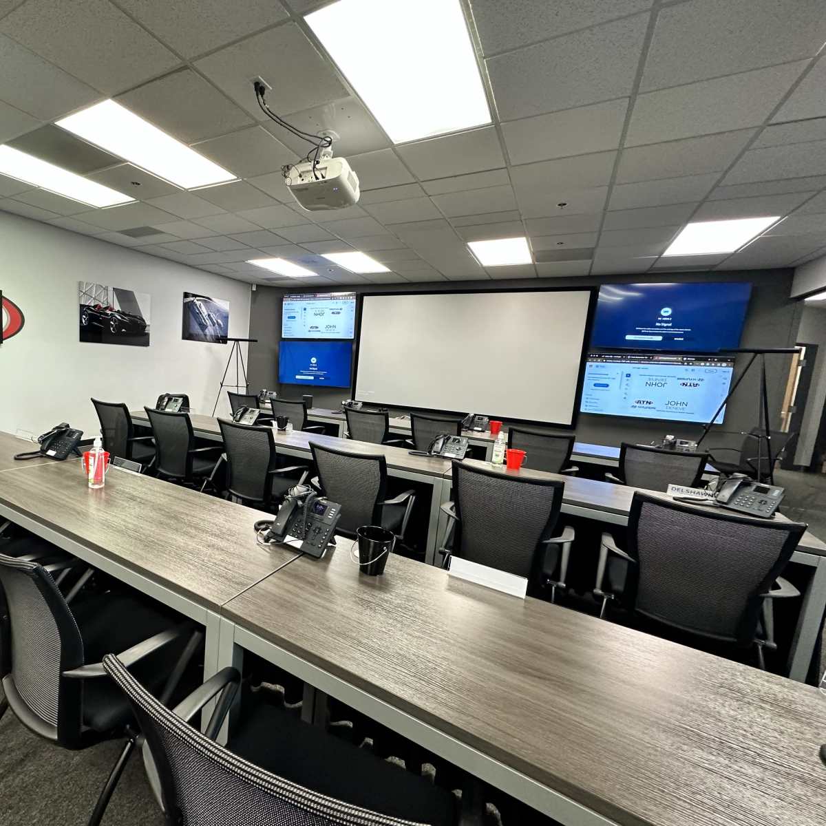 A modern conference room is equipped with multiple monitors, a projector, and desks arranged with telephones and chairs.