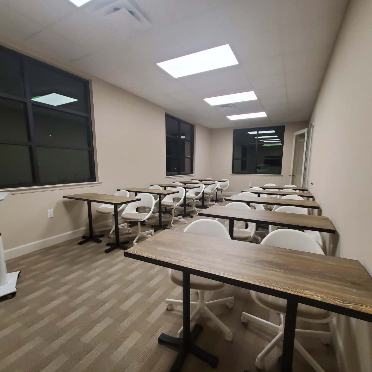 A classroom with several empty tables and chairs arranged in rows, illuminated by overhead lights and natural light from large windows.