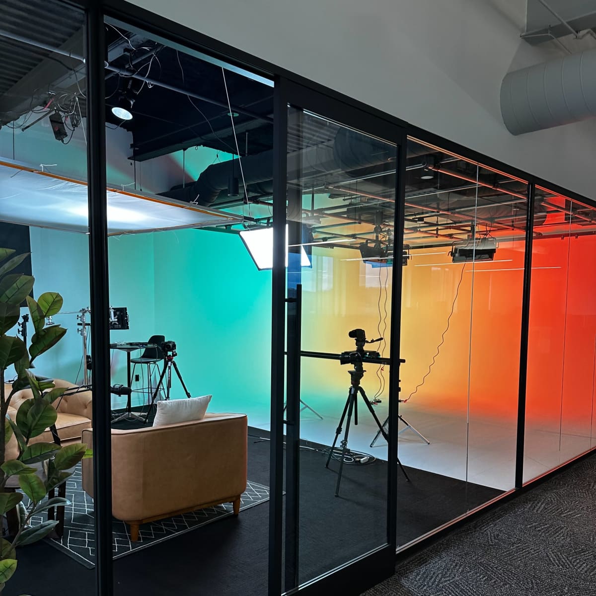 A modern studio space with a green to orange gradient wall, equipped with cameras and lights, visible through large glass walls.