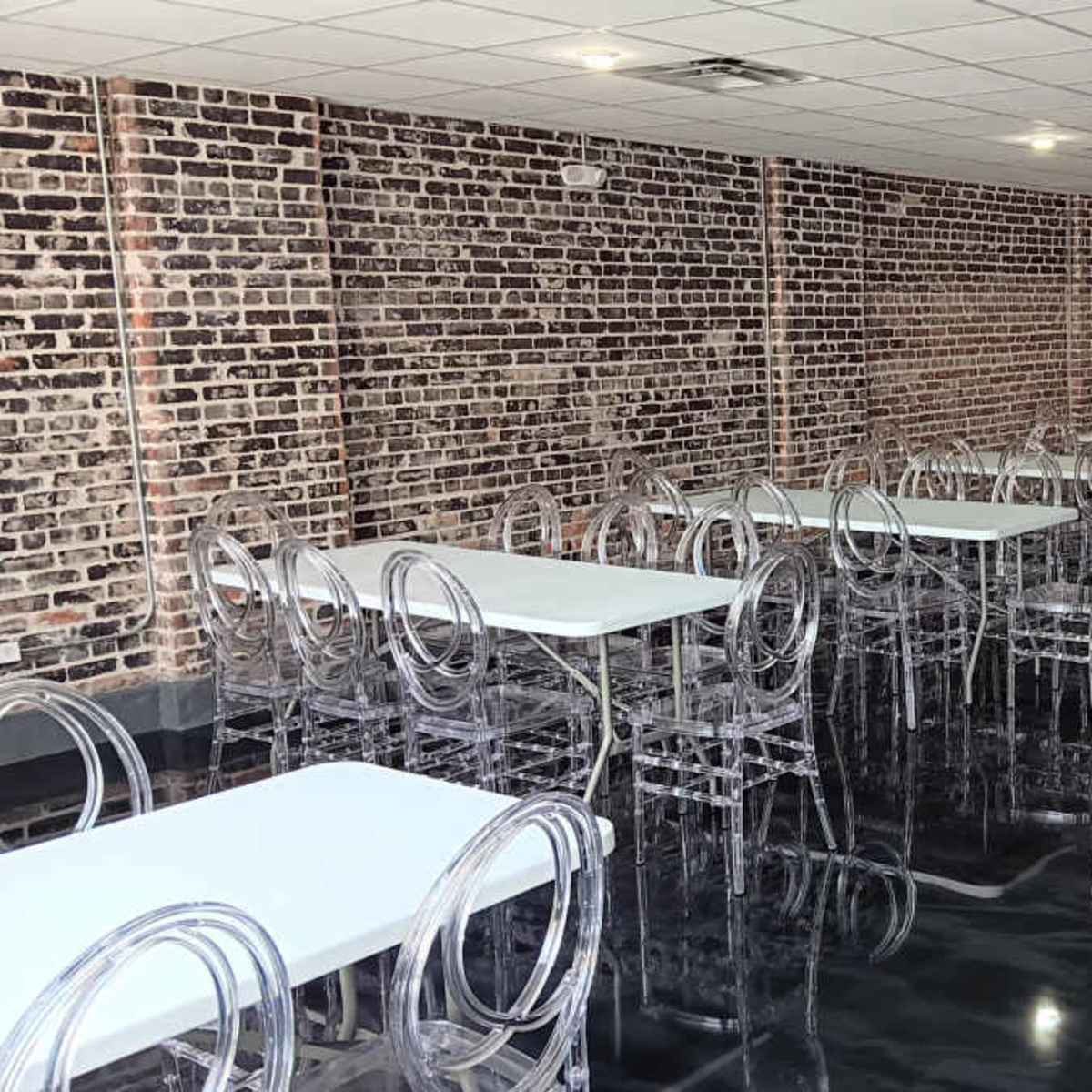 A spacious room with several tables and clear plastic chairs, featuring a brick wall and polished black flooring.