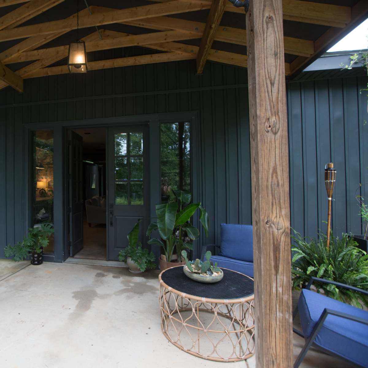 A covered porch with a hanging chair, a round coffee table, and potted plants in front of a dark-colored house.