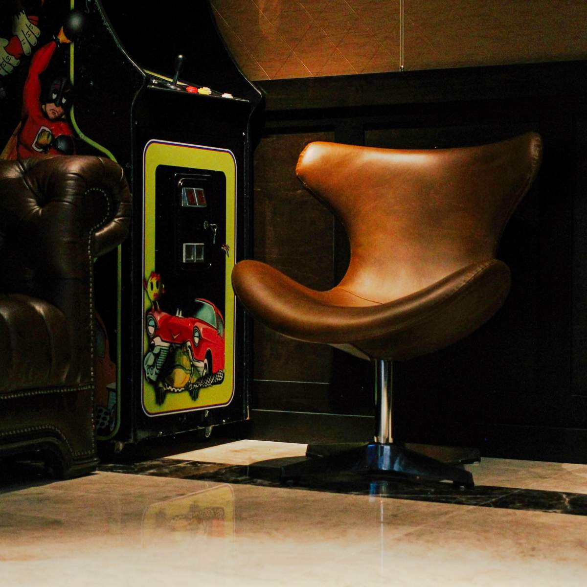 A modern brown chair beside a vintage arcade game in a dimly lit room with leather seating.