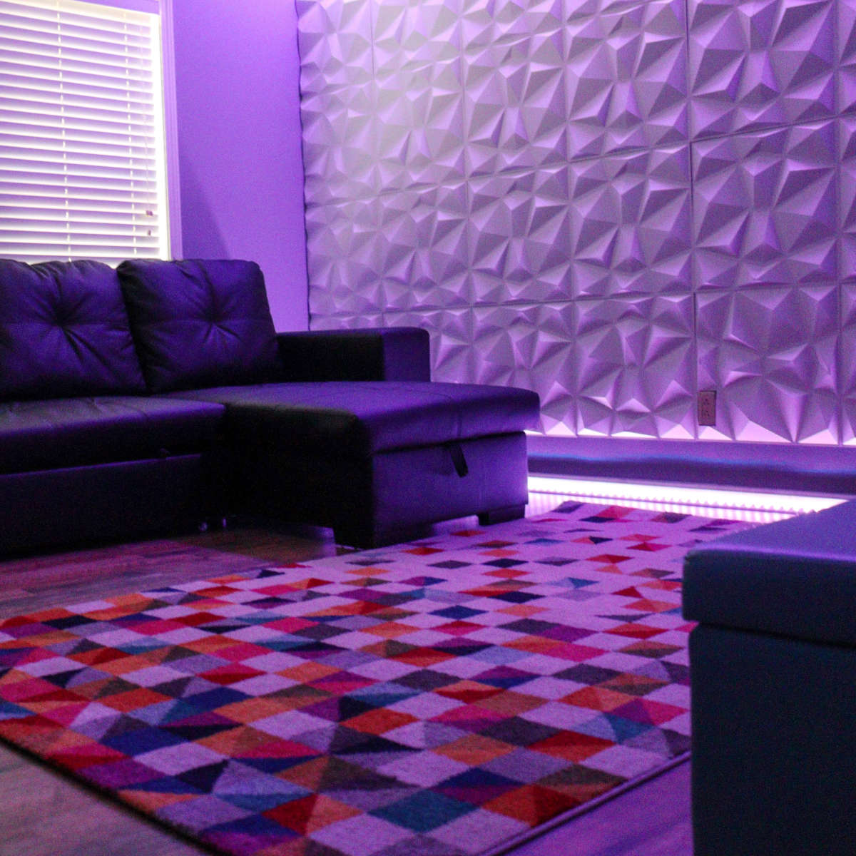 A black sofa and ottoman are arranged on a patterned rug in a room with purple lighting and textured wall panels.