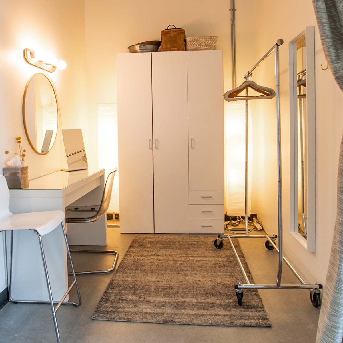 A small dressing room with a mirror, a chair, a clothing rack, and a storage cabinet against the wall, illuminated by soft lighting.