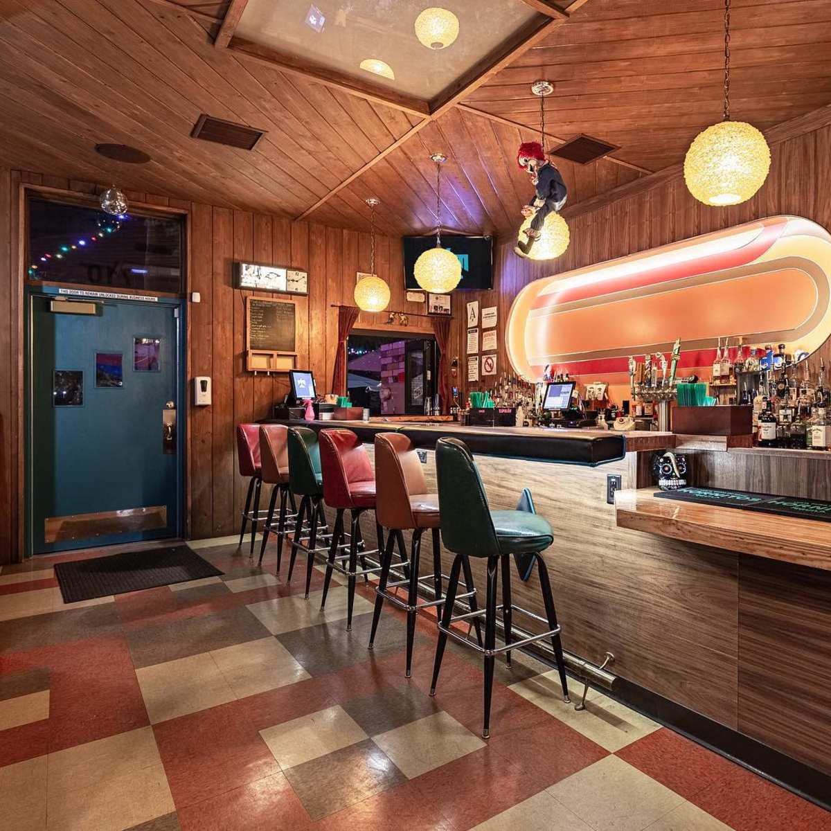 A bar area with a wooden interior, colorful stools, and a well-stocked bar illuminated by hanging pendant lights.