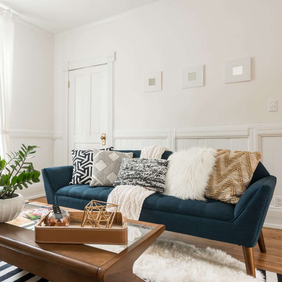 A modern living room features a blue sofa adorned with decorative pillows, a wooden coffee table, and houseplants, all set against white walls and natural light.