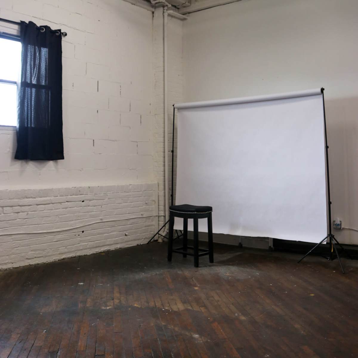 A black stool is positioned in front of a white backdrop in a well-lit, empty room with wooden floors and a window covered by a dark curtain.