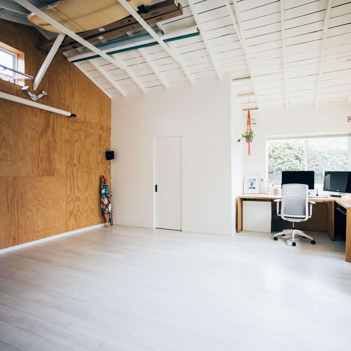 A spacious office features light wood paneling, two desks with computers, and minimal decor, illuminated by natural light from a large window.