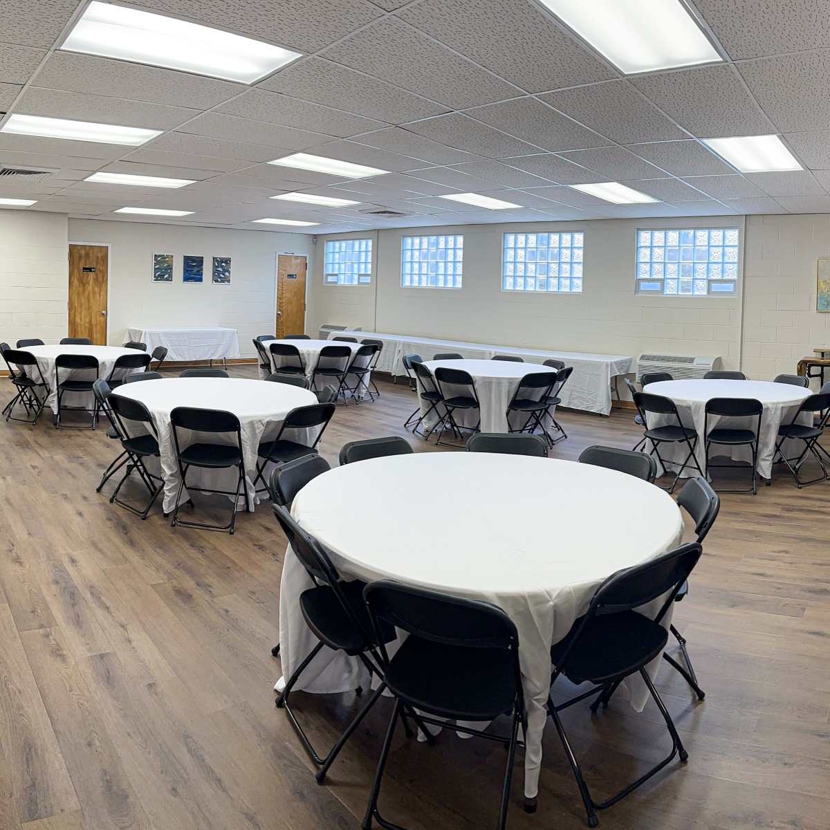 A large, well-lit room with multiple round tables covered in white tablecloths and black chairs arranged around them.