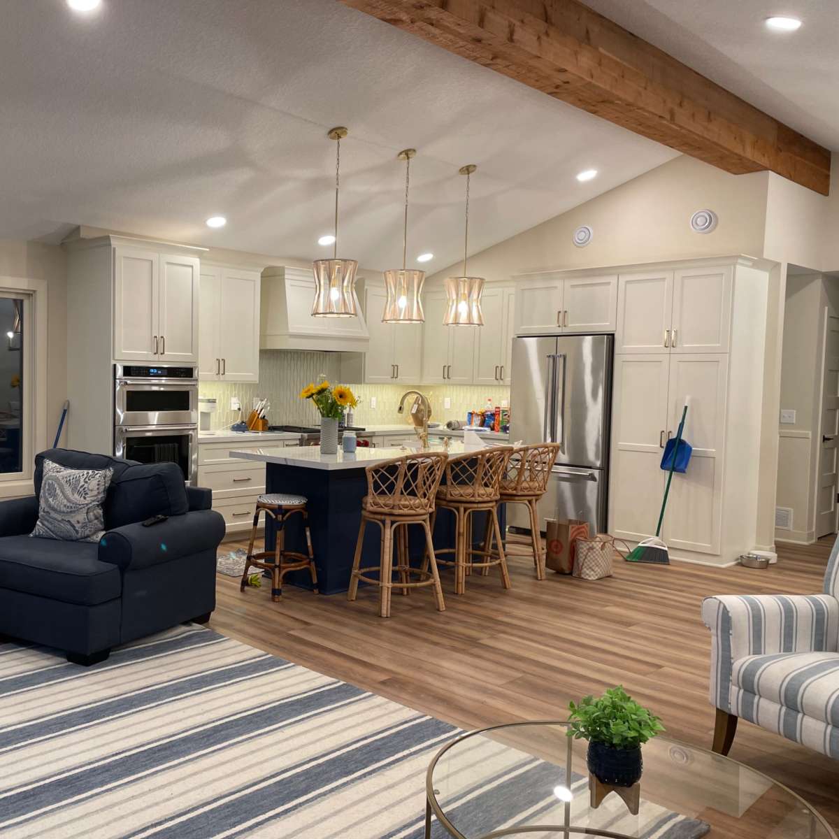 A modern kitchen and living area with light-colored cabinetry, four bar stools at the island, and a sectional sofa with a striped rug.