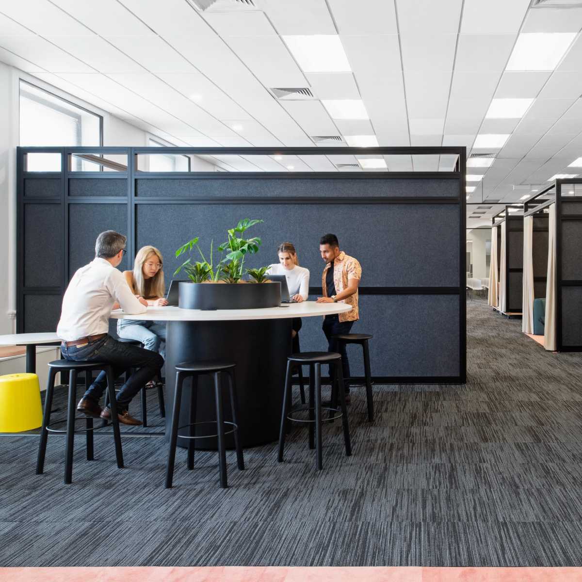 A modern office space features a group of people collaborating at a round table surrounded by partitioned workstations and a lounge area.