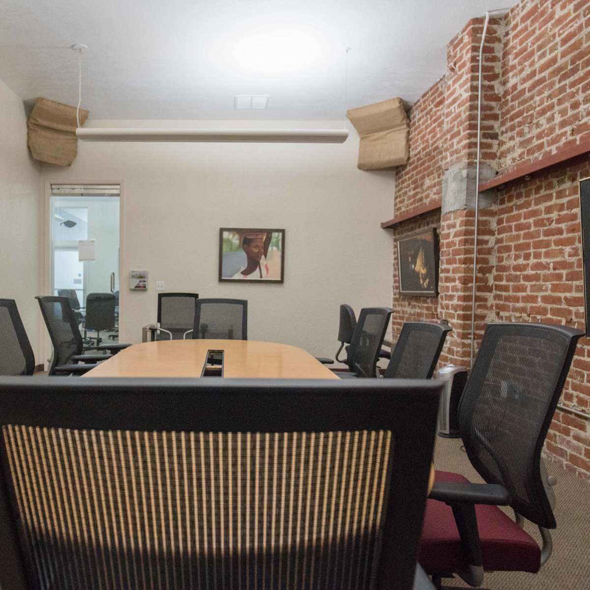 A conference room features a long table surrounded by ergonomic chairs, with exposed brick walls and artwork displayed.