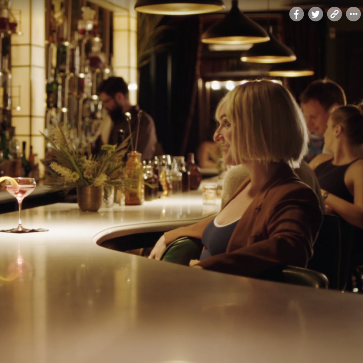 Two people are seated at a bar, with one man looking at a woman while a bartender prepares drinks in the background.