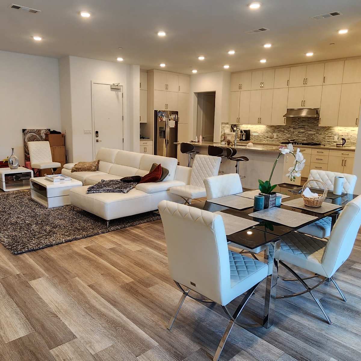 A modern living room features a white sectional sofa, a glass dining table with chairs, and an open-concept kitchen in the background.