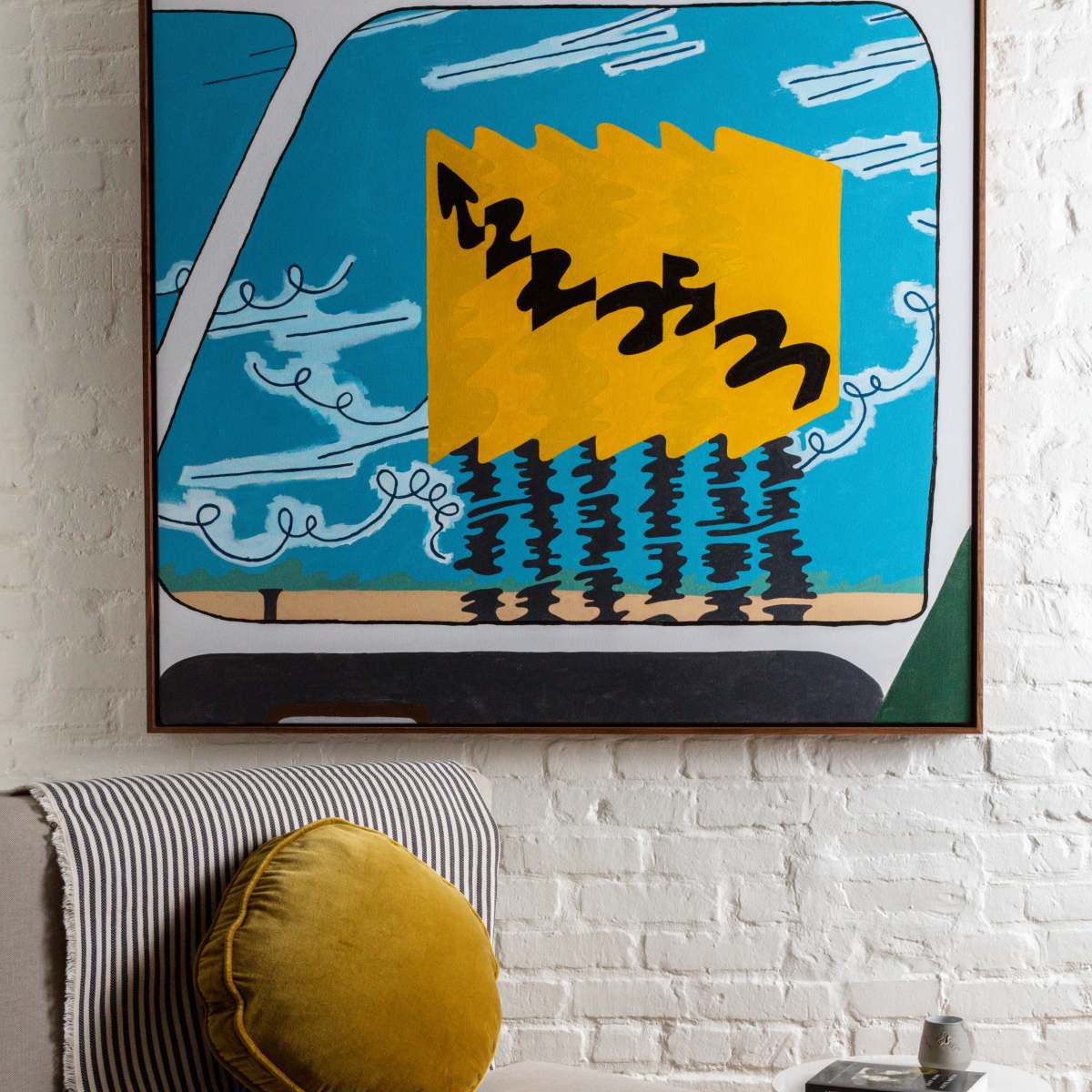 A framed abstract painting with bold colors and geometric shapes hanging above a light-colored sofa with a round yellow pillow.