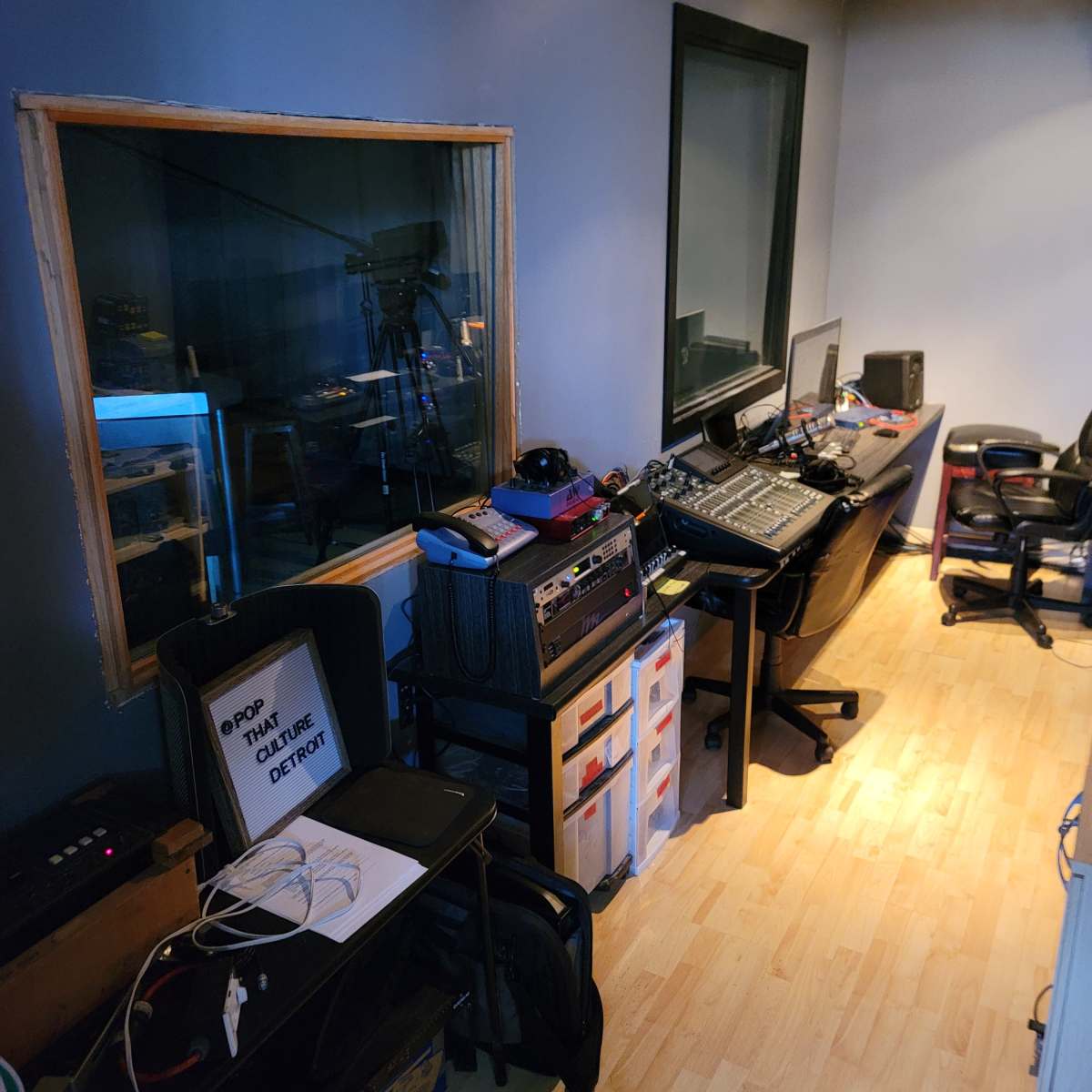 A recording studio with a control desk, various audio equipment, and a large window overlooking a sound booth.
