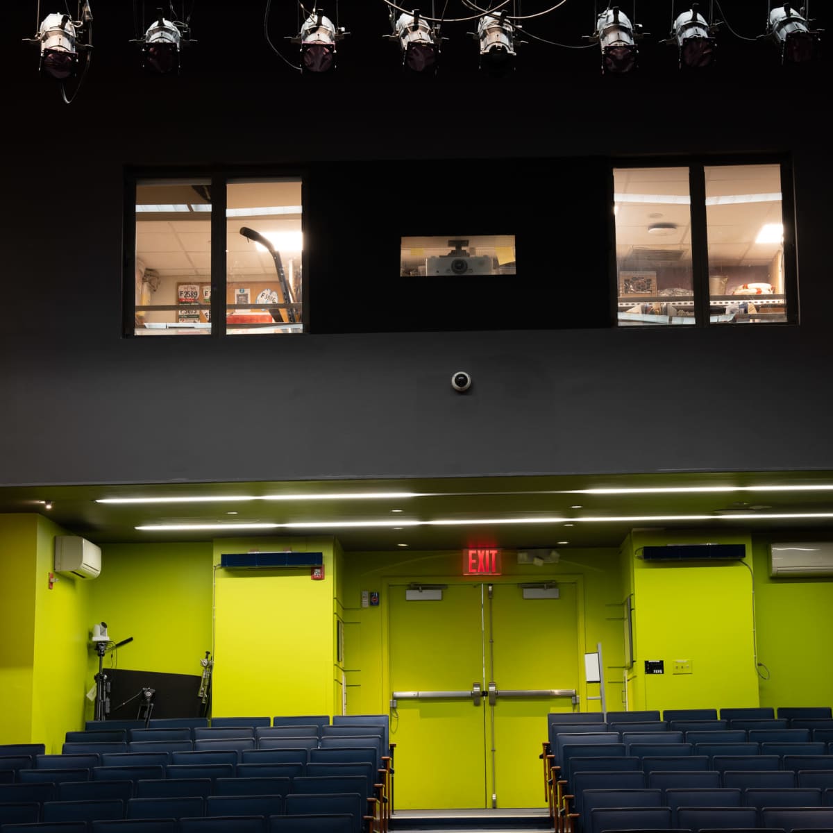 An empty auditorium with rows of blue seats and a green-lit stage area, featuring a control room window and emergency exit doors.