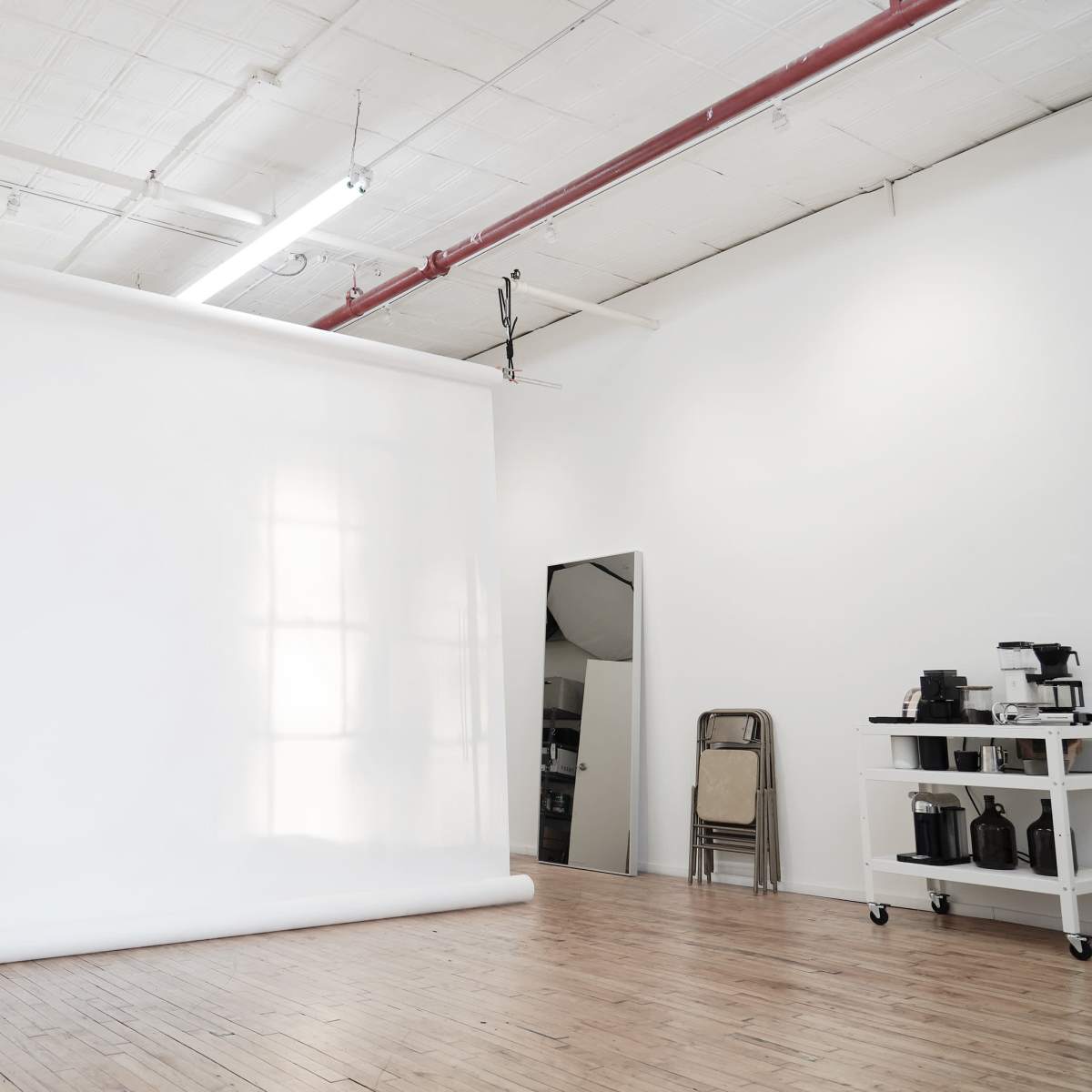 A spacious photography studio with a large white backdrop, wooden flooring, and a rolling chair beside a coffee station.