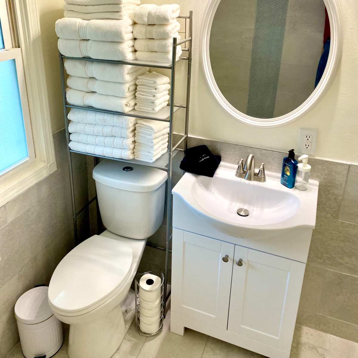 A neatly organized bathroom features stacks of white towels on a metal rack beside a toilet and a sink with a round mirror above it.