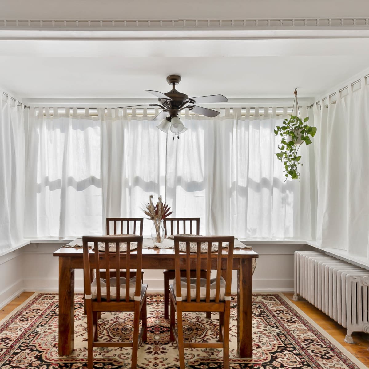 A dining area features a wooden table with four chairs, surrounded by large windows covered with sheer white curtains and a radiator against the wall.