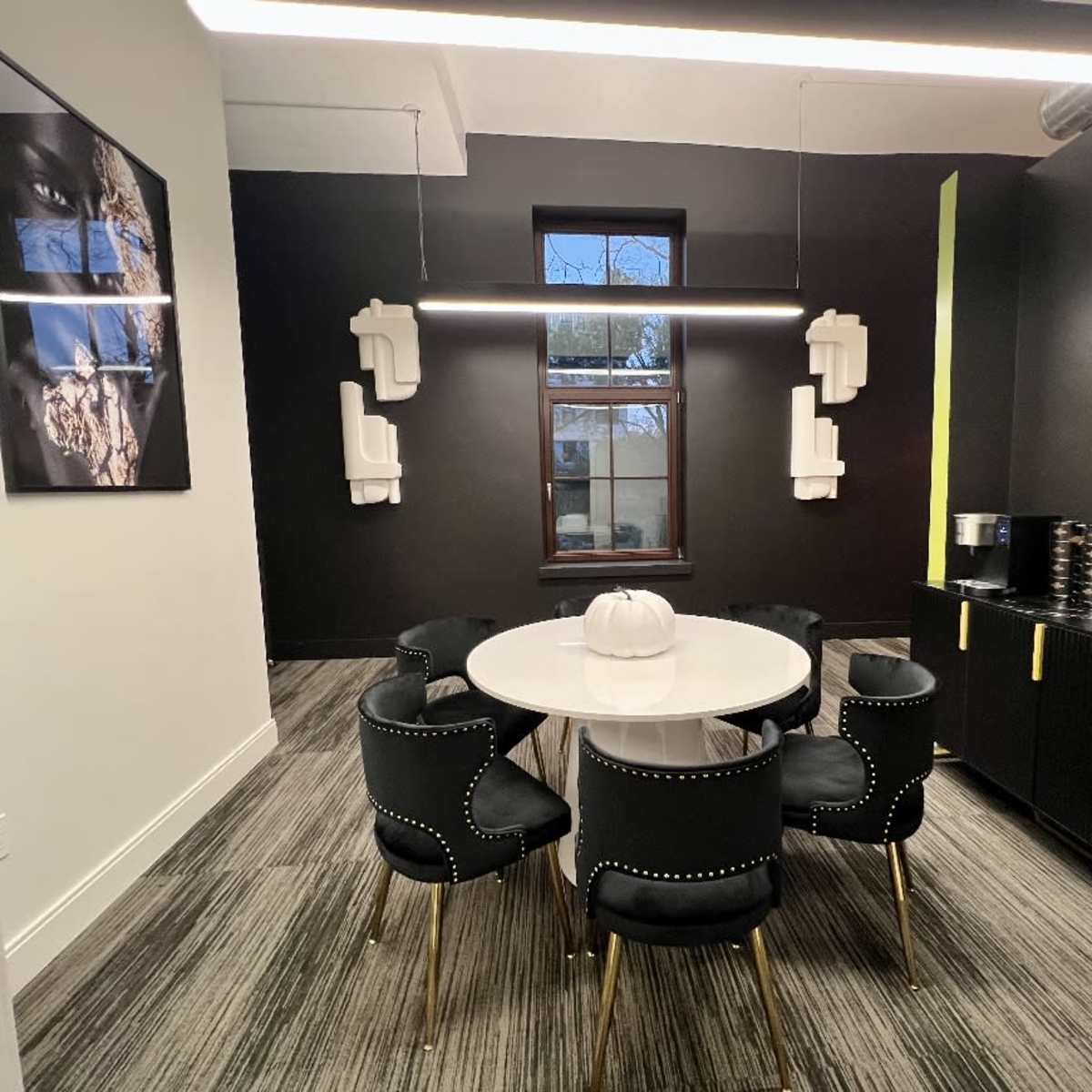 A modern meeting room features a circular white table surrounded by six black chairs with gold accents, along with a wall-mounted television and artwork.