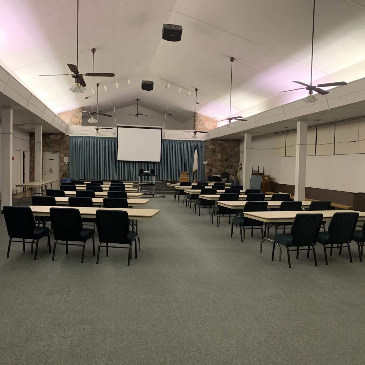 A multipurpose room set up with rows of tables and chairs facing a projector screen at the front.