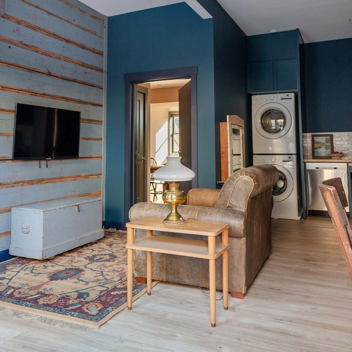 A modern living room with a brown sofa, a lamp on a wooden table, a television mounted on a textured wall, and laundry appliances visible in the background.