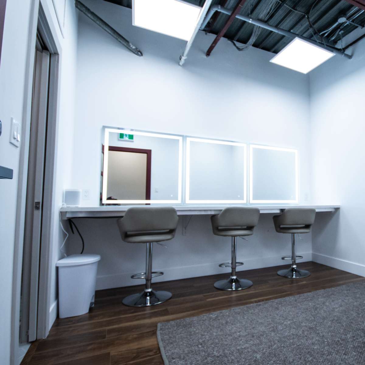 A minimalist makeup room featuring three bar stools at a long counter with three illuminated mirrors, a wastebasket, and a carpeted floor.