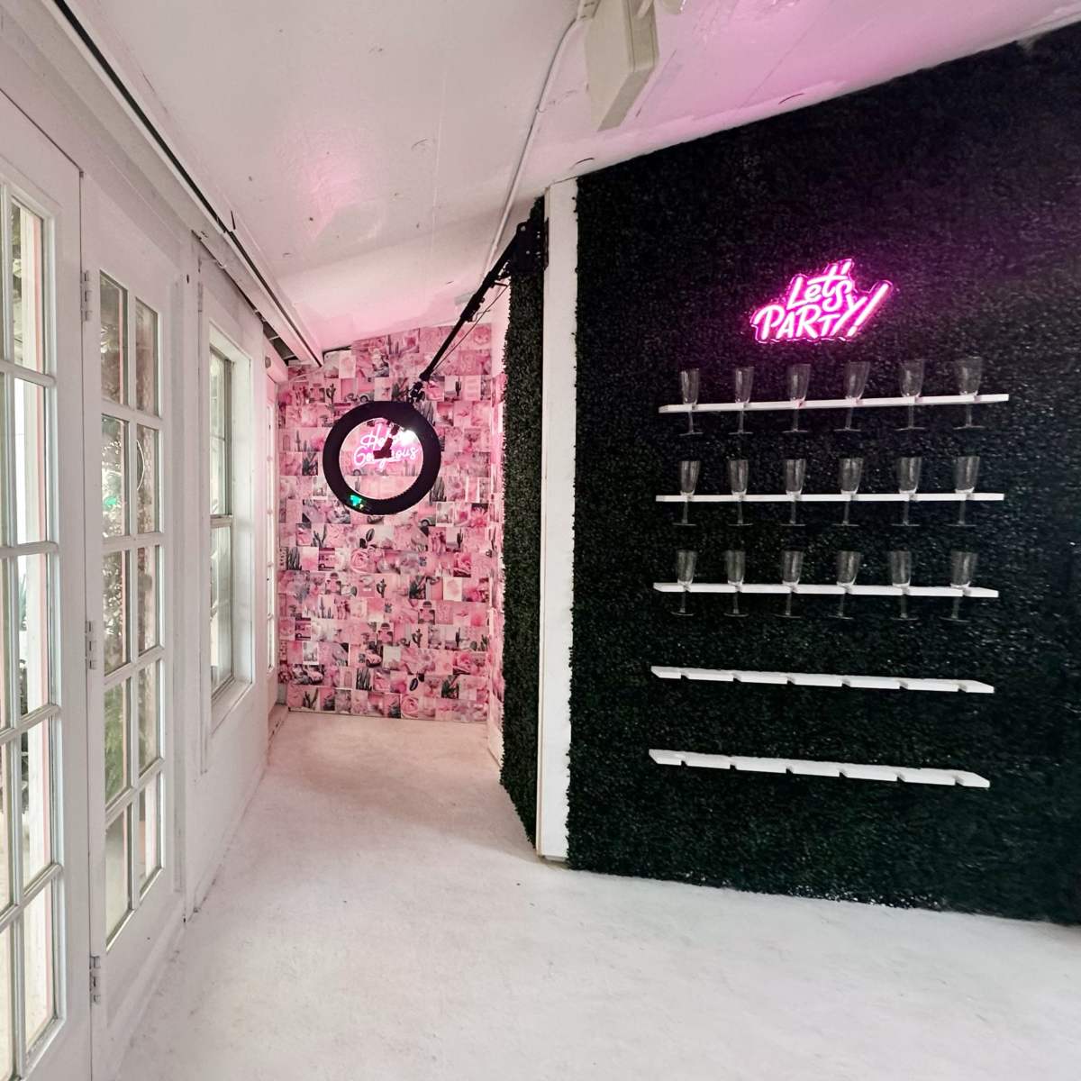A brightly lit hallway with glass doors leading to a wall decorated with pink and black photo collages and a neon sign that reads "Let's PARTY!" next to shelves displaying clear glasses.