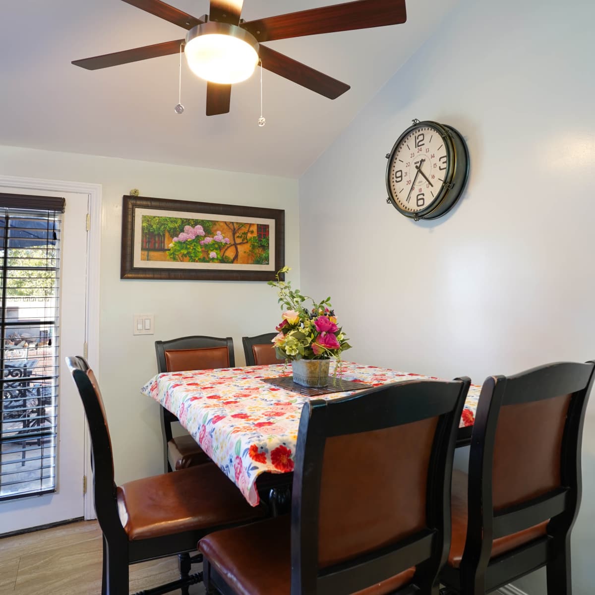 A dining area features a table covered with a floral tablecloth surrounded by high-backed chairs, with a ceiling fan and wall clock above.