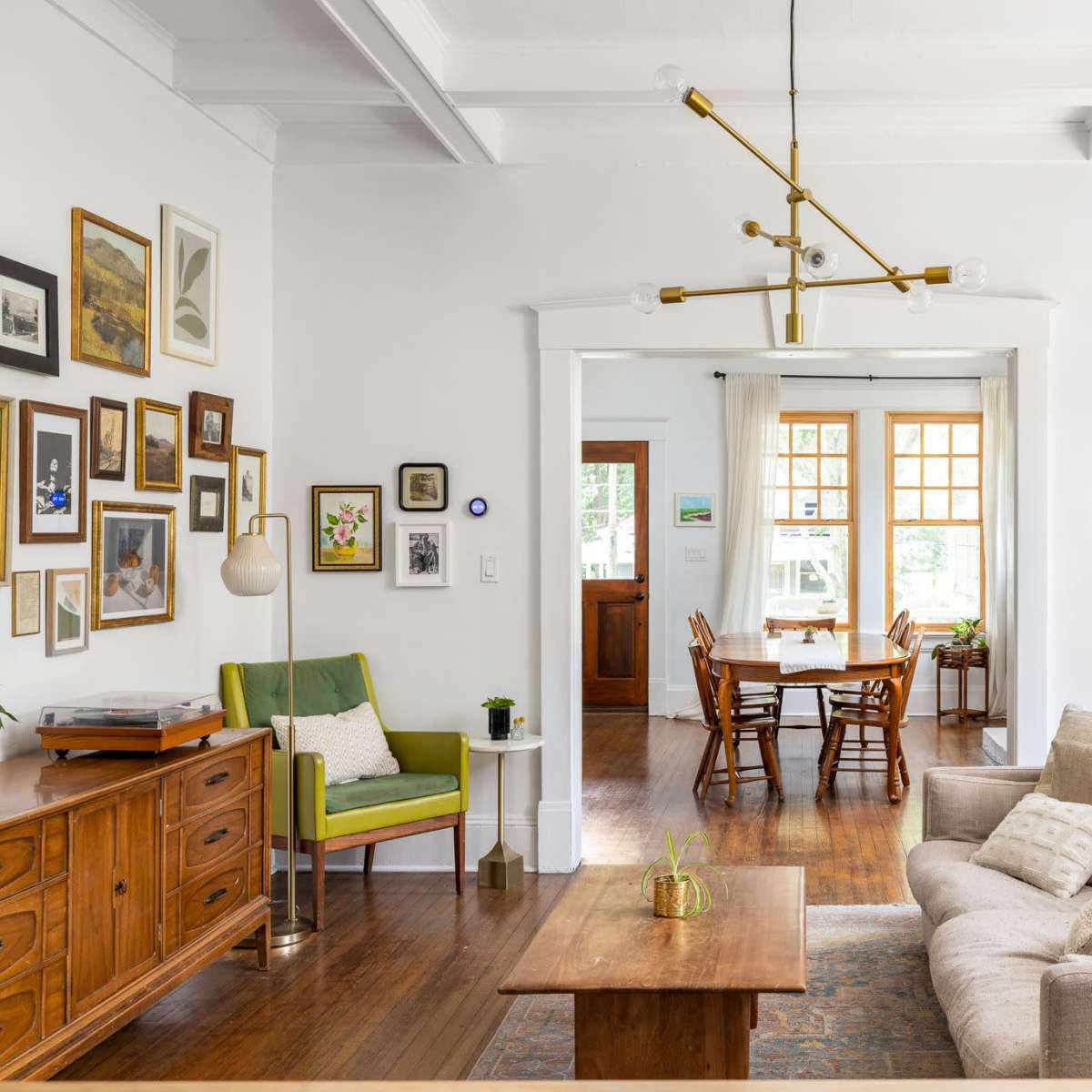A bright living room features a beige sofa, wooden furniture, a green accent chair, and a dining area visible through an archway.