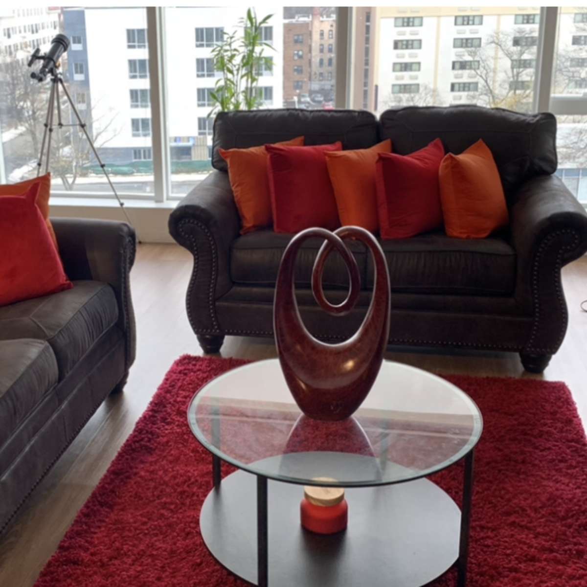 A modern living room features two dark sofas adorned with red and orange pillows, a glass coffee table with a decorative vase, and large windows displaying an urban view.