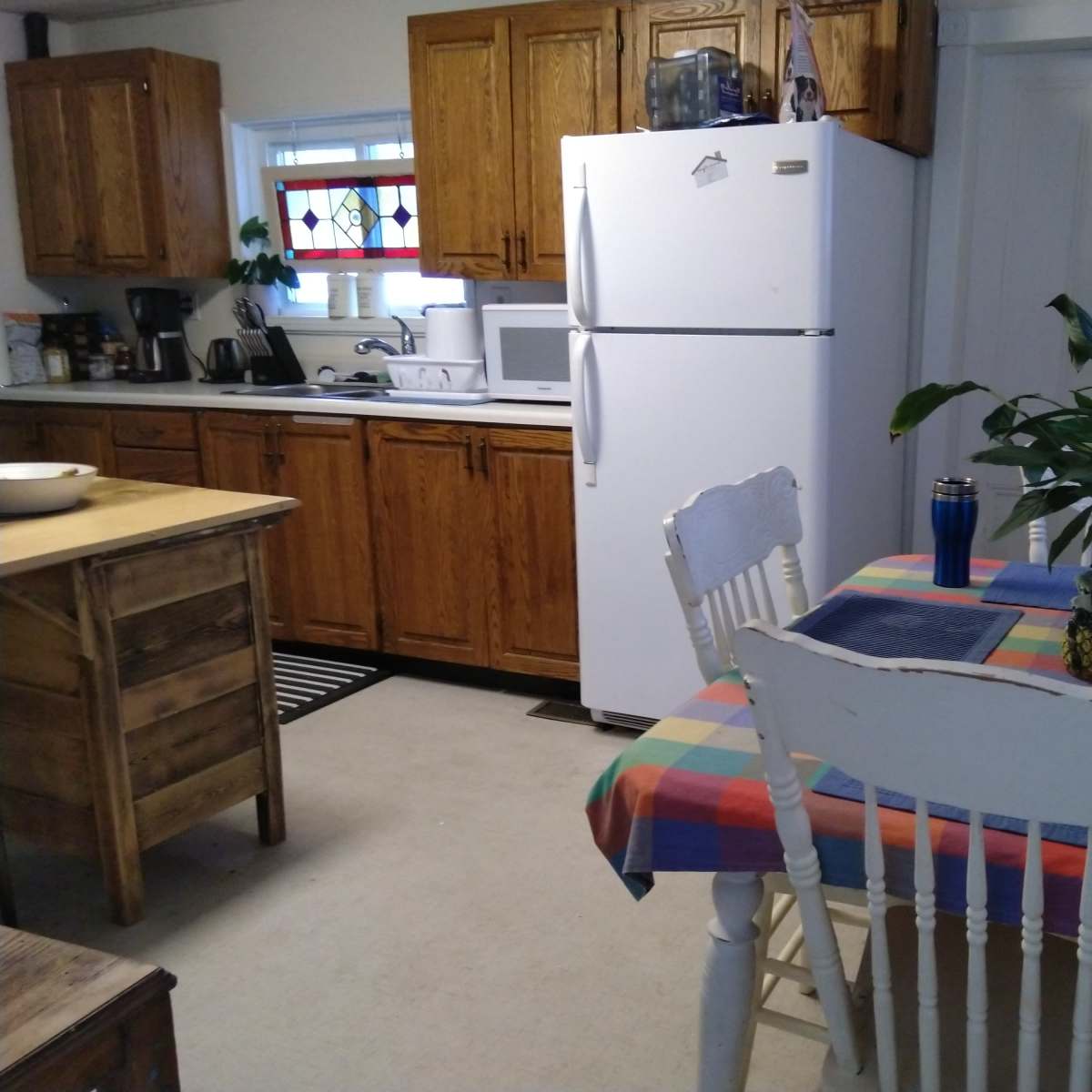 A cozy kitchen features wooden cabinets, a white refrigerator, a dining table with a colorful checkered cloth, and a potted plant.