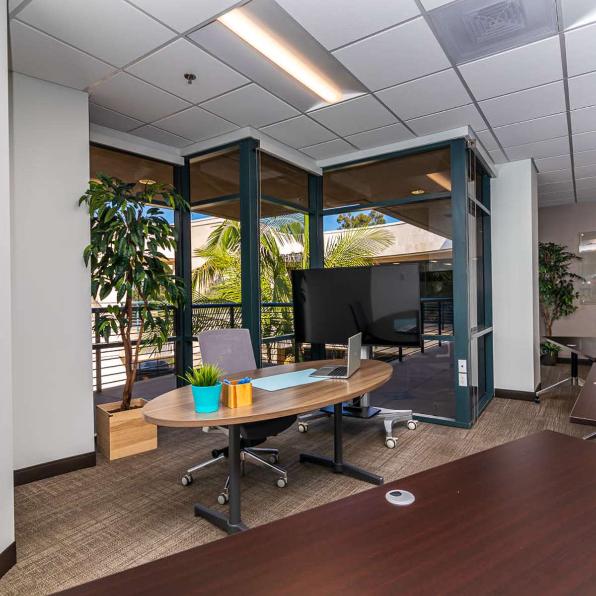 A modern office space with glass doors, a large screen on a stand, and multiple tables arranged for meetings, surrounded by greenery.