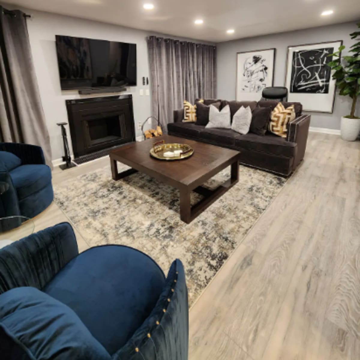 A modern living room featuring a dark sofa with multiple pillows, a wooden coffee table, two blue chairs, and a wall-mounted television above a fireplace.