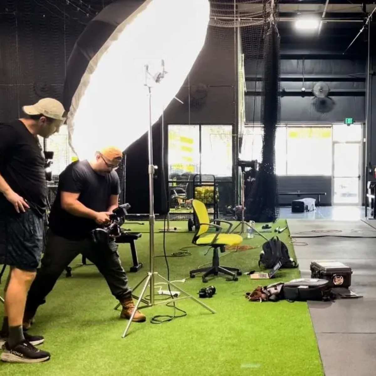 A photoshoot is taking place in a gym with a female model posing while two photographers adjust their equipment.